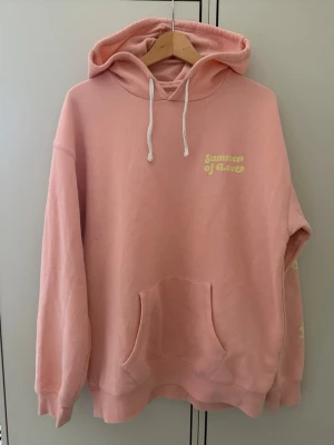Rosa hoodie från Billabong - Snygg rosa hoodie från Billabong med vit dragsko i huvan och trycket 'Summer of Love' i gult på bröstet. Mjuk insida och stor känguruficka framtill. Perfekt för chill dagar och streetstyle. 