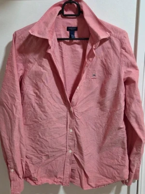 Gant Women Skjorta Ljusrosa Storlek S/36 - En klassisk och tidlös skjorta från Gant Women i en mycket klädsam ljusrosa nyans. Skjortan är i mycket fint skick och har den välkända kvalitet och passform som Gant är kända för. Observera: En knapp vid ena handleden är halv (se sista bilden), men då original-extraknappar medföljer på tvättlappen är detta enkelt åtgärdat för den som vill byta. I övrigt är skjortan i fint skick! Axlar: 39 cm Längd: 56 cm. Skick: Mycket fint skick (extraknappar medföljer)