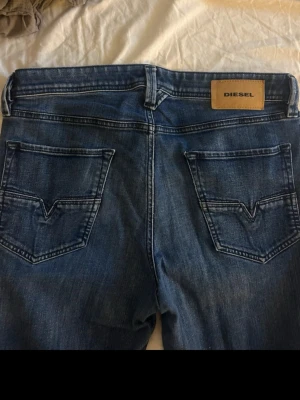 Blå jeans från Diesel med raka ben - Säljer ett par klassiska blå jeans från Diesel med raka ben och snygga slitningar. Jeansen har fem fickor, tydliga sömmar och en läderpatch med Diesel-loggan bak i midjan. Materialet är denim i bomull och passformen är normal. Perfekta för dig som gillar tidlös stil.