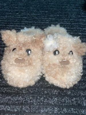 Fluffiga tofflor  - Supergulliga tofflor i beige och vitt med lockigt fluff och söta nalleansikten framtill. De har små öron, broderade detaljer och en mjuk, mysig känsla. Perfekta för att hålla fötterna varma hemma. De är knappt använda och är i storlek 33/34 men passar även större. De är unisex.