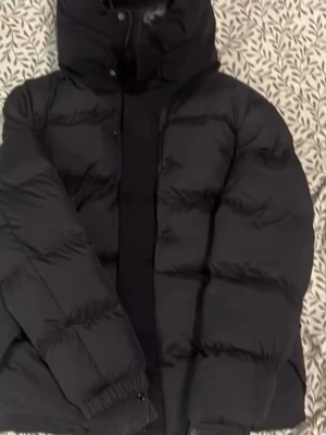 Svart pufferjacka med huva - Snygg svart pufferjacka med hög krage och stor huva. Jackan har breda kanaler, dragkedja och tryckknappar framtill samt elastiska muddar vid ärmsluten. Materialet är glansigt och ger en modern look. Perfekt för kalla dagar.