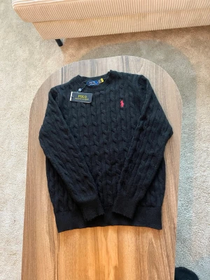 Svart kabelstickad tröja Polo Ralph Lauren - Svart kabelstickad tröja från Polo Ralph Lauren med klassisk design och röd broderad logga på bröstet.  • Storlek: enligt etikett (se bild) • Passform: Regular fit • Skick: Oanvänd – nyskick, med originaltag kvar • Färg: Svart • Detaljer: Kabelstickning, rund halsringning, ribbade muddar och nederkant, röd Polo-logga på bröstet  Helt oanvänd tröja i perfekt skick. En tidlös Polo-klassiker som funkar lika bra till vardag som mer uppklädda outfits.  Nypris: ca 1 700–1 900 kr 