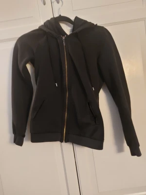 Svart hoodie med dragkedja - En svart hoodie med dragkedja framtill och två fickor. Modellen har huva med dragsko och ribbade muddar vid ärmslut och nederkant. Perfekt för en avslappnad och stilren look.