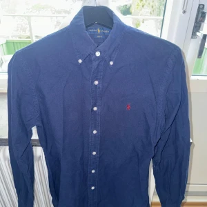 Mörkblå skjorta från Ralph Lauren - Klassisk mörkblå skjorta från Ralph Lauren med knappkrage och vita knappar. Skjortan har lång ärm och den ikoniska röda logotypen broderad på bröstet. Tillverkad i bomull och passar perfekt till en stilren look.