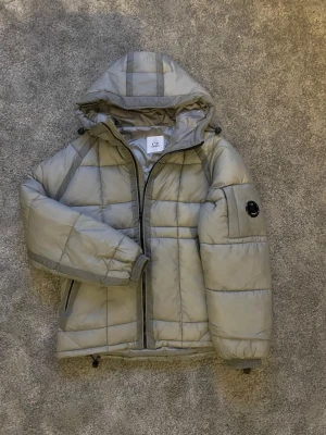 Grå pufferjacka från C.P. Company - Snygg grå pufferjacka från C.P. Company med huva och dragkedja framtill. Jackan har ett emblem på ärmen, två sidofickor med dragkedja och justerbar nederkant. Quiltad design och vadderad för extra värme. Perfekt för kalla dagar.