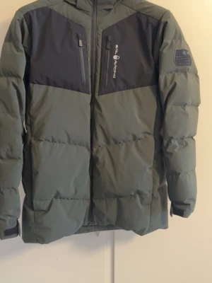 Grön pufferjacka från Sail Racing - Säljer en grön pufferjacka från Sail Racing med svarta detaljer på bröstet och två bröstfickor med dragkedja. Jackan har patch på ärmen med texten 'Sail Antarctica Expedition' och är perfekt för kalla vinterdagar. Snygg, sportig look och riktigt varm.