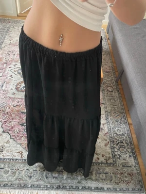 Svart långkjol med volang - Säljer en svart långkjol med bred resår i midjan och volangdetaljer. Kjolen har en luftig och flowy passform, perfekt för dig som gillar en avslappnad men ändå stilren look.