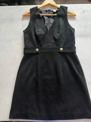 Svart ärmlös klänning med bälte - Stilren svart klänning i mjukt material med v-ringning och bred midjedetalj med två guldfärgade knappar. Klänningen är ärmlös, har en figurnära passform och dragkedja i ryggen. Perfekt för dig som gillar minimalistisk och chic stil.