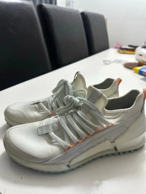 Vita sneakers från ECCO - Snygga vita sneakers från ECCO med bred tåbox och minimalistisk design. Skorna har ljusgrå snören, diskreta orange detaljer och en flexibel sula. Tillverkade i syntetmaterial och textil för en lätt och luftig känsla.