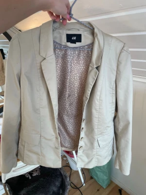 Beige kavaj från H&M - Stilren beige kavaj från H&M med klassisk krage och knappstängning framtill. Kavajen har figurnära passform. Perfekt att matcha till olika outfits för en snygg och modern look.