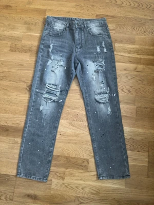 Grå slitna jeans med färgstänk - Säljer ett par grå jeans med raka ben, slitningar och hål på framsidan samt vita färgstänk över hela byxorna. Jeansen har klassisk femficksmodell och en cool, avslappnad streetstil. Perfekta för dig som gillar en edgy look.