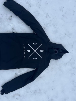 Svart hoodiejacka från Dope - Svart hoodiejacka från Dope med stor vit logga och text på bröstet. Jackan har huva med dragsko, stor magficka och långa ärmar. Materialet är vattenavvisande och passar perfekt för utomhusaktiviteter och streetwear.