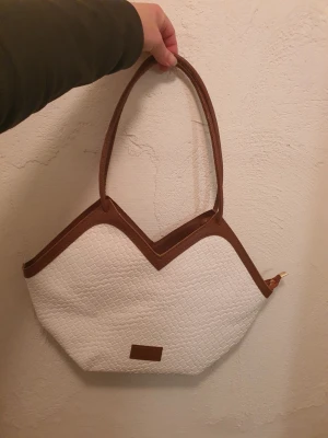 Vit handväska med bruna detaljer - Snygg handväska i vitt med bruna detaljer från Fashion & Bags. Väskan har ett unikt sicksack-mönster upptill och är gjord i ett flätat konstläder. Praktiska handtag och en liten brun logopatch framtill. Perfekt för dig som gillar trendiga accessoarer. Helt nu.