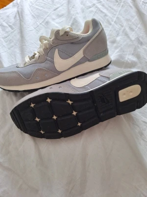 Nike gråvita sneakers low - Snygga sneakers från Nike i grått och vitt med klassisk swoosh på sidan. Ovandelen är i mesh och mocka för en sportig look och skön känsla. Yttersulan är svart med bra grepp och mellansulan är vit. Perfekta för dig som gillar stilrena och bekväma skor.