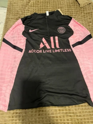 PSG träningströja svart/rosa Nike - Snygg långärmad träningströja från PSG i svart med rosa detaljer och loggor. Tröjan har halv dragkedja, klubbmärke på bröstet och texten 'Accor Live Limitless' samt 'Visit Rwanda' på ryggen. Tillverkad i ett lätt och funktionellt material.