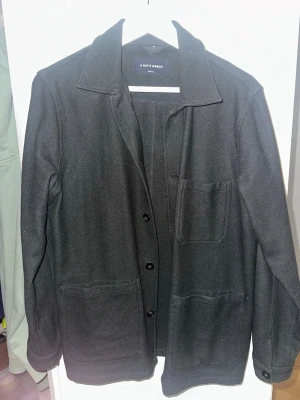 Svart overshirt från A Day's March - Stilren svart overshirt från A Day's March med klassisk krage, knäppning framtill och tre praktiska fickor. Tillverkad i ett mjukt, tjockare tyg som ger en clean look. Perfekt lager-på-lager-plagg för dig som gillar minimalistisk stil.    Använd en gång. 