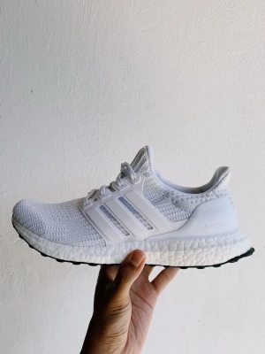 Vita Adidas Ultraboost sneakers - Säljer ett par vita Adidas Ultraboost sneakers med stickad ovandel i mesh, klassiska tre ränder på sidan och Boost-sula för extra komfort. Skorna har snörning och låg profil som ger en clean och sportig look. Perfekta för dig som gillar stilrena och bekväma sneakers.