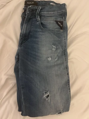 Blå slitna jeans från Replay - Snygga ljusblå jeans från Replay med slitningar och hål på framsidan. Klassisk femficksmodell med logga på fickan och bakficka med broderad detalj. Perfekta för en avslappnad och trendig look.