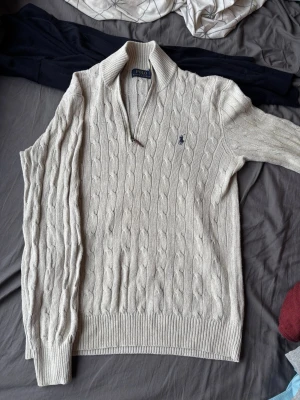 Halfzip Polo Ralph Lauren - Kabelstickad beige tröja från Polo Ralph Lauren med halv dragkedja och klassisk krage. Tröjan har lång ärm och den ikoniska blå loggan broderad på bröstet. Perfekt för dig som gillar tidlös och stilren design.