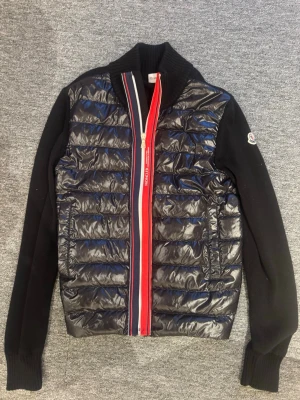 Svart cardiganjacka från Moncler - Snygg svart cardiganjacka från Moncler med quiltad, glansig front och stickade ärmar. Dragkedjan har röda och vita detaljer och jackan har hög krage samt Moncler-logga på ärmen. Perfekt för dig som vill ha en sportig men stilren look. Storleken är 14 år men det motsvarar xxs.