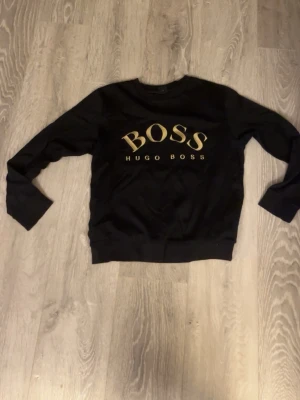 Svart sweatshirt från Hugo Boss - Svart sweatshirt från Hugo Boss med stort guldigt BOSS-tryck på bröstet. Klassisk rund halsringning och långa ärmar. Perfekt för en stilren och avslappnad look.