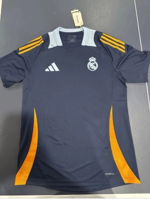 Real Madrid träningströja Adidas blå - Mörkblå Real Madrid träningströja från Adidas med ljusblå detaljer vid halsen och orangea inslag på sidorna och axlarna. Tröjan har klubbmärke och Adidas-logga på bröstet samt Aeroready-material som andas. Perfekt för fotbollsträning.