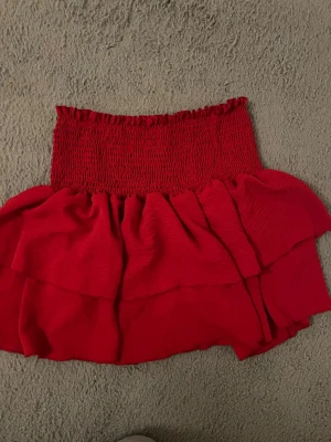 Röd volangkjol med smock - Säljer en kort röd kjol med smock upptill och dubbla volanger. Kjolen har en ungdomlig och trendig look, perfekt för dig som gillar att sticka ut. Materialet känns mjukt och lätt, och den elastiska midjan gör den bekväm att bära.