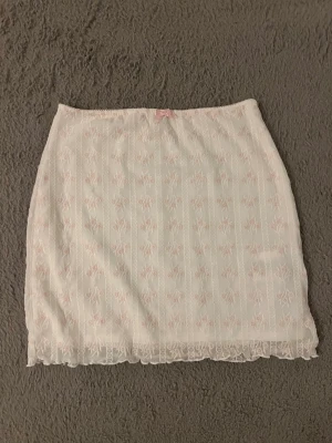 Vit spetskjol med rosett - Söt vit kortkjol med spetsdetaljer och en liten rosa rosett framtill. Kjolen har ett diskret blommigt mönster och är gjord i ett lätt material som passar perfekt för varma dagar. Spetsen längs nederkanten ger en romantisk touch. Blir lågmidjad på mig som brukar ha S men den är storlek L