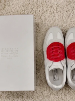 Maison Margiela vita sneakers med rött - Maison Margiela sneakers i vitt läder och mocka med unika röda detaljer över snörningen. Klassisk låg modell med vit sula och stilren design. Perfekta för dig som vill sticka ut med en exklusiv touch.