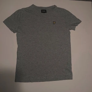 Grå t-shirt från Lyle & Scott - Snygg grå t-shirt från Lyle & Scott med klassisk passform och gul broderad logga på bröstet. Priset kan diskuteras.