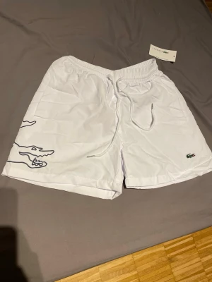 Vita badbyxor från Lacoste - Snygga vita badbyxor från Lacoste med elastisk midja och dragsko. Stora krokodiltryck på både fram- och baksida samt klassisk Lacoste-logga framtill. Lätta och snabbtorkande, perfekta för stranden eller poolen. De är mycket små i storlek.
