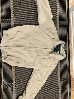 Beige vindjacka från Polo Ralph Lauren - Stilren beige vindjacka från Polo Ralph Lauren med dragkedja framtill och två sidofickor. Jackan har elastiska muddar och en liten mörkblå logga broderad på bröstet. Insidan är fodrad med blått rutigt tyg för extra stilpoäng.