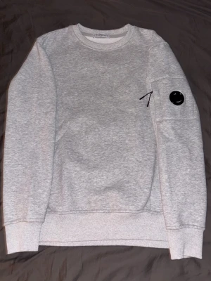 Grå sweatshirt från C.P. Company - Snygg grå sweatshirt från C.P. Company med rund halsringning och ribbade muddar. Tröjan har en ikonisk svart linsdetalj på armen och en praktisk ficka med dragkedja. Perfekt för en avslappnad och trendig look.