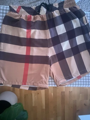 Burberyy Shorts/Badbyxor S - Säljer ett par klassiska rutiga shorts från Burberry med beige, svart, vit och röd färg. Shortsen har elastisk midja med snörning och är tillverkade i ett lätt syntetmaterial. Perfekta för sommaren och har den ikoniska Burberry-looken.