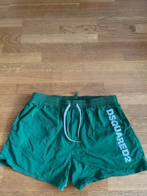 Gröna badbyxor från Dsquared2 - Snygga gröna badbyxor från Dsquared2 med vitt tryck längs benet och vita dragsnören i midjan. Klassisk kort modell med elastisk midja och sidofickor. Perfekta för stranden eller poolen.