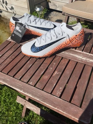 Nike Mercurial fotbollsskor grå/orange - Säljer ett par helt nya utan box Nike Mercurial fotbollsskor i grått med svart och blå swoosh samt orange mönster längs sidorna. Skorna har snörning och en färgglad yttersula med dobbar för bra grepp på planen. Perfekta för dig som vill sticka ut på fotbollsplanen.