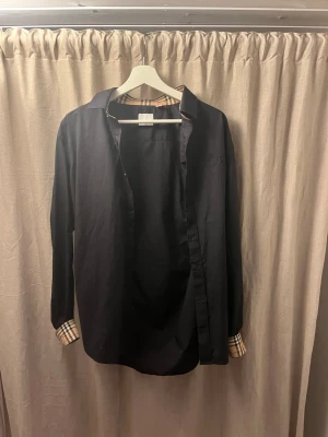 Svart Burberry skjorta med rutiga detaljer - Snygg svart skjorta från Burberry med klassiska rutiga detaljer på insidan av kragen och ärmsluten. Skjortan har knappar framtill och är långärmad, perfekt för en stilren look. Står att det är strl XL men den passar mig som brukar ha M