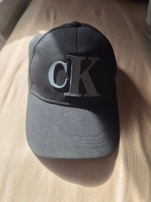 Svart keps från Calvin Klein - Snygg svart keps från Calvin Klein med stort CK-logotyp framtill och justerbart spänne bak i metall. Klassisk böjd skärm och stilren design, perfekt för dig som gillar streetwear och enkel stil. Tillverkad i slitstarkt tyg.