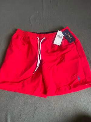 Röda badbyxor från Polo Ralph Lauren - Snygga röda badbyxor från Polo Ralph Lauren med vit dragsko i midjan och klassisk blå logga broderad på benet. Tillverkade i snabbtorkande material och har elastisk midja för skön passform. Perfekta för stranden eller poolen.