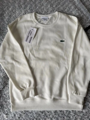 Vit sweatshirt från Lacoste - Klassisk vit sweatshirt från Lacoste med rund halsringning och det ikoniska gröna krokodilmärket broderat på bröstet. Tröjan har ribbade muddar vid ärmslut och nederkant. Perfekt för en clean och stilren look.