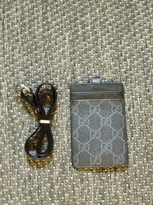 Grå Gucci korthållare med rem - Snygg grå korthållare från Gucci med klassiskt GG-mönster. Plånboken har ett fack med genomskinlig framsida för ID eller kort och kommer med en matchande svart rem som kan fästas i metallögla. Tillverkad i slitstarkt material med exklusiv känsla.