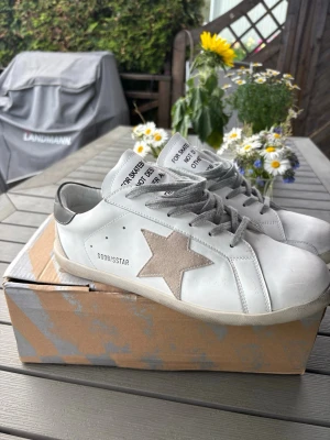 Vita Golden Goose sneakers med stjärna - Säljer ett par vita Golden Goose sneakers med grå snören och beige mockastjärna på sidan. Skorna har en klassisk låg siluett och detaljer i läder och mocka. Perfekta för dig som gillar en avslappnad men trendig stil.
