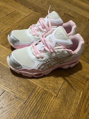 ASICS sneakers i vitt och rosa - Snygga ASICS sneakers i vitt och ljusrosa med silvriga detaljer. Skorna har mesh- och läderpaneler, rosa snören och en chunky sula. Perfekta för dig som gillar sportig och trendig stil.