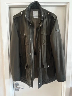 Grön fieldjacka från j.lindeberg - En mörkgrön feildjacket från j. Lindeberg. Perfekt för lite kyligare kvällar. Jackan har en liten defekt på den västra fickan men det påverkar inte användningen. Jackan har också en infällbar luva och även justerbara snören vid midjan. Hör av er vid minsta lilla fråga 😀