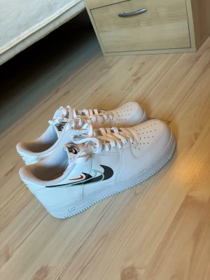 Nike Air Force 1 vita sneakers - Fräscha vita Nike Air Force 1 sneakers med flera färgade swoosh-loggor på sidan i svart, grått och orange. Klassisk låg modell med perforerad tå, vita skosnören och sula i gummi. Perfekta för dig som vill ha en clean men ändå unik look.