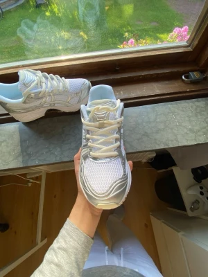 Vita och silver Asics sneakers - Snygga Asics sneakers i vitt och silver med mesh- och syntetdetaljer. Skorna har en chunky sula och klassisk Asics-logga på sidan. Perfekta för dig som gillar sportig och trendig stil. Lätta och bekväma med ventilerande material.