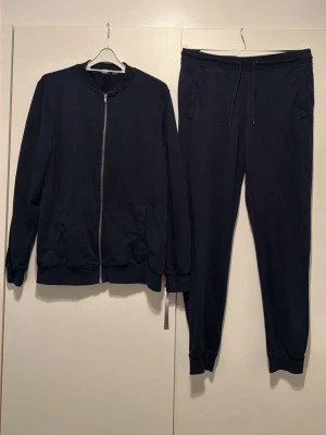 Mörkblått set med dragkedja - Snyggt mörkblått set med långärmad tröja med dragkedja och matchande byxor med dragsko i midjan. Tröjan har två fickor framtill och ribbade muddar. Setet är gjort i mjukt bomullsliknande material och har en avslappnad passform.