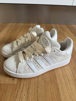 Adidas original Campus 00’s sneakers i beige mocka - Snygga beige sneakers från Adidas i mjuk mocka med klassiska vita ränder på sidorna och Adidas-logga på plösen. Skorna har breda, krämvita skosnören och en låg silhuett som ger en clean och stilren look. Perfekta för dig som gillar streetwear och enkel design. Endast använda/provade en gång. 
