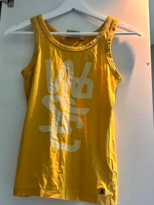 Gul tanktop från WESC - Snygg gul tanktop från WESC med stort vitt tryck framtill och logga nertill. Har en klassisk ärmlös design med rund halsringning. Perfekt för sommardagar eller som ett lager under skjortan.