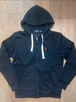 Svart hoodie från Polo Ralph Lauren - Svart hoodie med dragkedja från Polo Ralph Lauren. Klassisk design med vit dragsko i huvan och röd broderad logga på bröstet. Mjuk bomullsmix, känguruficka framtill och ribbade muddar vid ärmar och nederkant. Perfekt för en chill och stilren look.